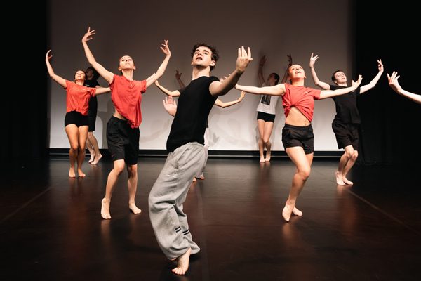 Dancers in a triangle formation raise their arms out to the side and gaze up, they appear to have their weight on one leg while the other leg is bent and the heel of the foot is lifted.