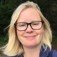 A woman with shoulder length blonde hair is smiling at the camera. She is wearing glasses.
