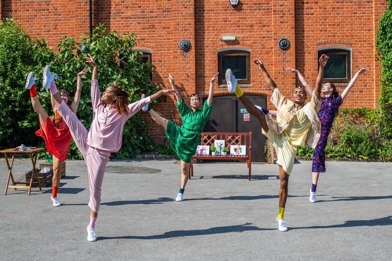 Dancers in bright clothing outside red brick building are all raised on one foot while they high kick the other leg with arms raised.