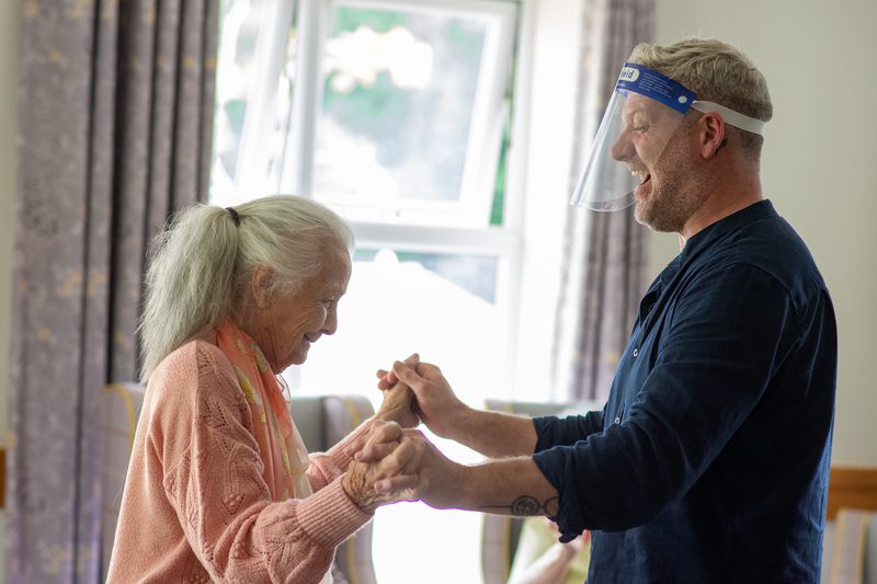 A new adventures dancer and a care home resident laughing as they hold hands to dance.