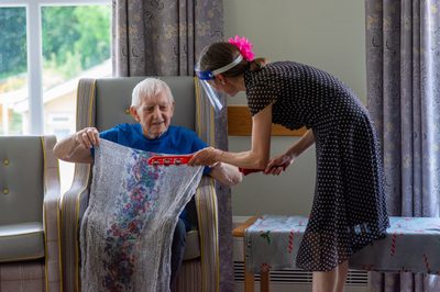 A smiling care home resident holds a floral scarf and converses with a dancer holding a tambourine.