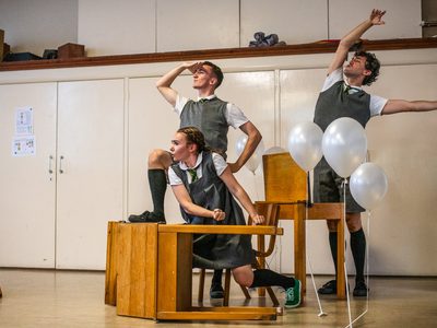 Three performers in grey school uniforms. Desks are arranged around them as they take on poses as if on a boat. There are white balloons hovering around them.