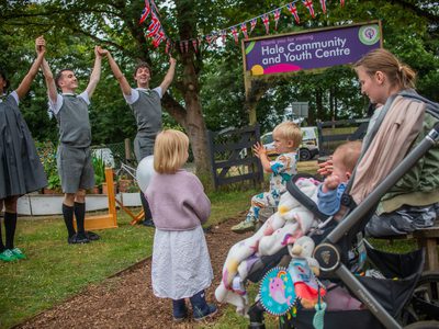 Three performers in grey school uniforms in a grassy setting take each other’s hands and lift them up as a young family watch.