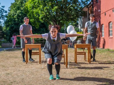 A performer in grey school uniform gazes directly forward, she has her knees bent and arms wide.