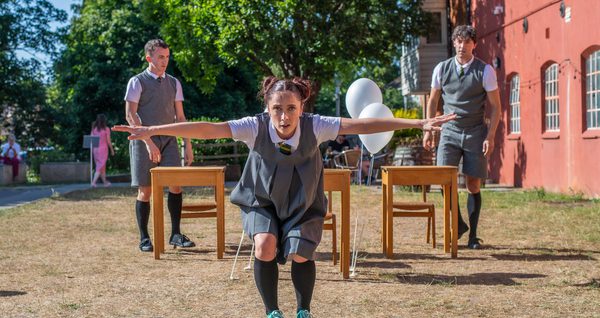 A performer in grey school uniform gazes directly forward, she has her knees bent and arms wide.