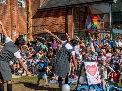 Performers in grey school uniforms make aeroplanes shapes, the audience does the same. They are in a grassy setting with a brick red building in the background.