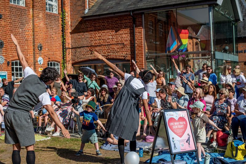 Performers in grey school uniforms make aeroplanes shapes, the audience does the same. They are in a grassy setting with a brick red building in the background.
