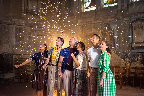Group of men and women in 50s clothing with arms around each other gazing up at sparkles as they fall from the ceiling