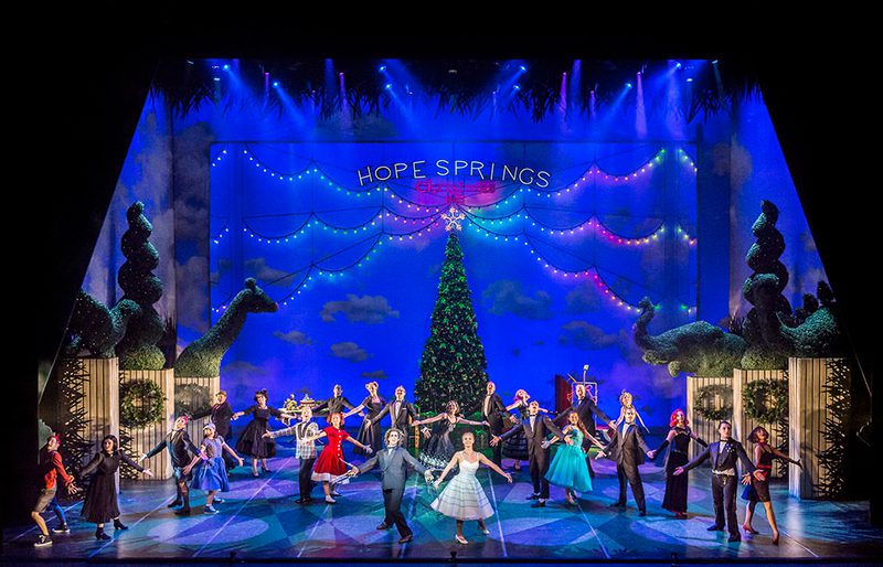 Edward and the towns people gather to celebrate Christmas. A large Christmas tree is in the background. Everyone stands with their arms spread out smiling.