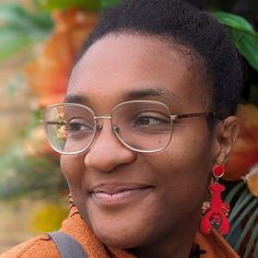 Elaine, a black woman in her thirties is smiling, staring to the left. Her afro-texture hair is in a fluffy bun and she’s wearing thin-rimmed glasses, red earrings in the shape of lobsters, and an orange coat. Behind her is a pillar decorated in many varieties of yellow, pink, and red flowers.