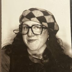 A black and white photo of Bronagh, a white woman in her twenties. She is wearing wide frame glasses and a checkered beret hat and smiling, looking into the camera lens.