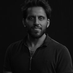 South Asian man, with wavy hair and a medium to short beard. Wearing a 1/4 zip, short sleeve polo shirt