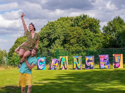 On green grass, a male in yellow shorts lifts a female in a black and yellow dress. She has her hand raised. Large letters behind them on the grass say CHANGE!