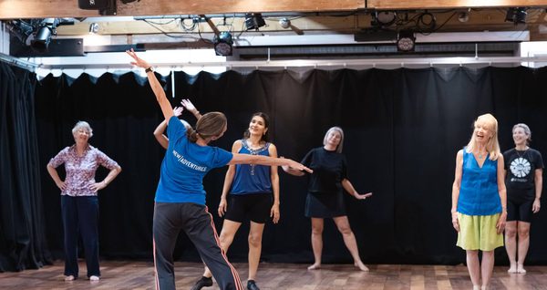 An instructor in a blue t-shirt is demonstrating a movement in a dance class. She is reaching her arms out at her sides, and the participants watch.
