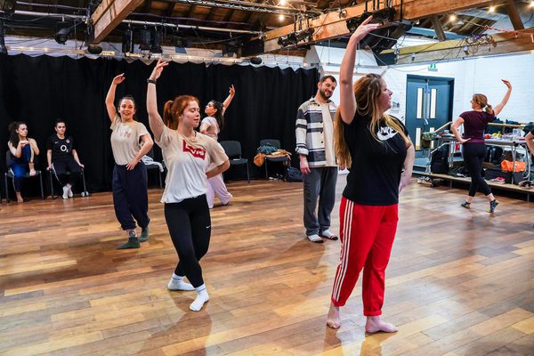 Several Overture participants stand in fourth position with one arm raised. One participant looks on with their head tilted to their left