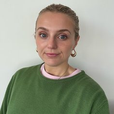 White female with fair hair pulled back off her face, she is wearing hooped earrings and a green jumper