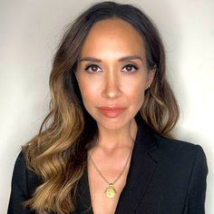 A woman with long wavy brown hair is looking at the camera. She is wearing a black v-neck top and a necklace.