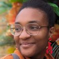 Elaine, a black woman in her thirties is smiling, staring to the left. Her afro-texture hair is in a fluffy bun and she’s wearing thin-rimmed glasses, red earrings in the shape of lobsters, and an orange coat. Behind her is a pillar decorated in many varieties of yellow, pink, and red flowers.
