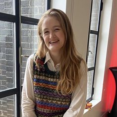 Person smiling, wearing a colorful knitted vest and a white shirt, seated indoors near a window with black frames.
