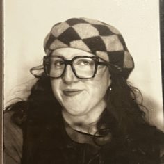 A black and white photo of Bronagh, a white woman in her twenties. She is wearing wide frame glasses and a checkered beret hat and smiling, looking into the camera lens.