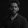 South Asian man, with wavy hair and a medium to short beard. Wearing a 1/4 zip, short sleeve polo shirt