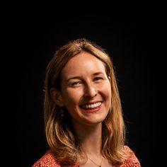 white woman with brown mid-length hair, wearing a red top and lipstick, smiling to the camera