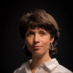 White female with freckles, hazel eyes and short brown hair, wearing hooped earrings and a crisp white shirt.