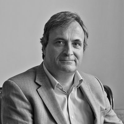 Black and white headshot of a man wearing a suit, sat down and smiling to camera.