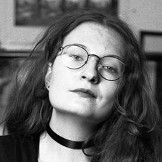 Black and white headshot of a person with long dark hair and round, dark glasses looking to camera.