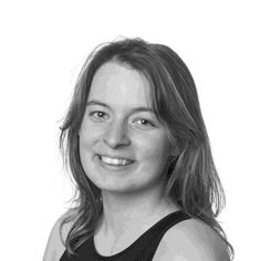 A white woman in her early 20s with long hair wearing a dark vest top, looking over her left shoulder, smiling towards the camera.