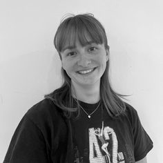 A white British woman, in her early 20's, with straight shoulder-length hair and a fringe, wearing a black 42nd Street Musical t-shirt, smiling at the camera.