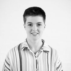 An upper-body photo of Katie, a white woman in her 20's with short ginger hair and brown eyes against a white background. She is wearing a stripy shirt with shiny buttons and is smiling towards the camera.