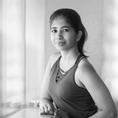 A South Asian woman in her late 20's, with hair tied back into a ponytail, wearing a dark top, smiling at the camera.