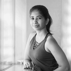 A South Asian woman in her late 20's, with hair tied back into a ponytail, wearing a dark top, smiling at the camera.