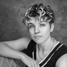 A white person in their early twenties, with short curly wavy blonde hair and one knee up with their right arm resting on it, gently smiling at the camera.