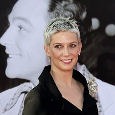 A woman with short blonde hair is standing with her head turned towards the camera, she is smiling. She is wearing a black, collared v-neck top.