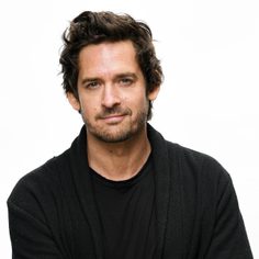 A man with dark coiffed hair is standing with his arms crossed looking at the camera. He is wearing a black t-shirt and a black cardigan.