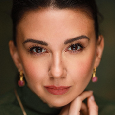 A woman is framed in a close-up and is looking at the camera. She has dark hair which is pulled up, and is wearing a dark green top and earrings. He hand is rested on her chin.