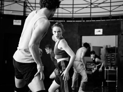 Rehearsal image of dancers facing off with their partners, their hands drawing up the outside of their thighs and an intense focus is in their eyes