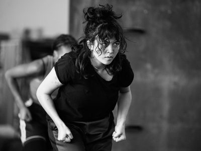 Rehearsal image of a dancer leaning forward with clenched fists by their hips and an intense expression on their face