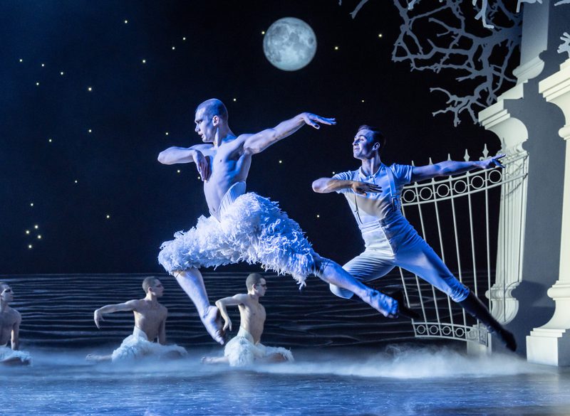 Two dancers are performing a dynamic jump on stage, set against a night-themed backdrop featuring a moon and stars, and silhouetted trees. Other dancers can be seen in the background, enhancing the atmospheric, setting of the lake.