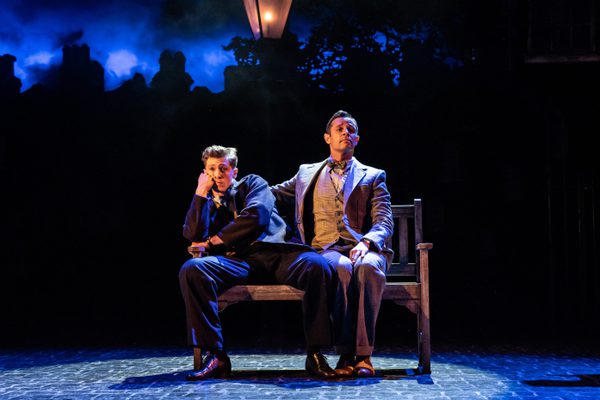 Two actors on stage in a theatrical production, sitting closely on a bench with a dimly lit, foggy cityscape background. Both are dressed formally, one in a dark suit and the other in a grey suit, portraying a scene of contemplation.