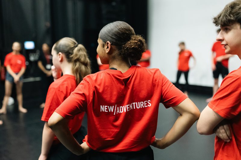 Dancers in red t shirts with New Adventures branding stand facing away from the camera into a busy room