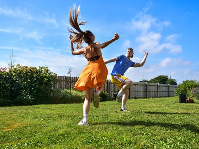 Female with long hair flying up in a ponytail and wearing an orange dress leaps towards a male in a blue t shirt and cap who has his front knee and arm lifted in glee