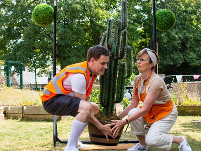 A comedy scene – two performers in orange lift a heavy cactus.