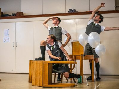 Three performers in grey school uniforms. Desks are arranged around them as they take on poses as if on a boat. There are white balloons hovering around them.