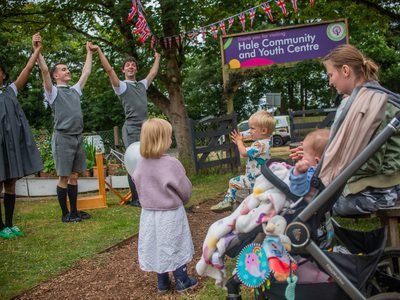 Three performers in grey school uniforms in a grassy setting take each other’s hands and lift them up as a young family watch.