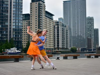 Two dancers in a classic partner dance pose with back hands clenched together, arms lifted, each has a leg outstretched, toes touching and they gaze down to their toes. There is a cityscape in the background.