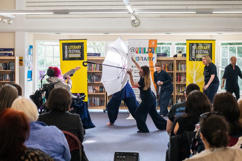 backs of heads watching a performance in a bright library space, performer hold large white umbrella and other performers reach towards the umbrella