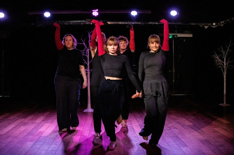 Flock of performers move forward as a group all dressed in black except for one arm which is red and raised like the long neck of a swan